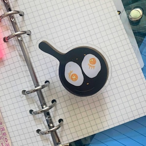 May include: A clear sticker featuring a cartoon illustration of a frying pan with two fried eggs. The eggs have orange yolks and white egg whites, with small decorative details. The pan is black with a smiling face.