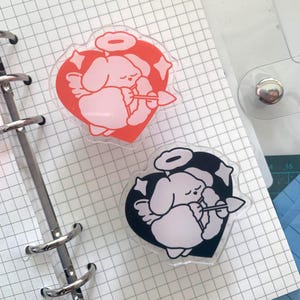 May include: Two heart-shaped stickers. One features a red heart with a pink angel bunny, the other a black heart with a white angel bunny. Both have halos, wings, and star accents, with a bow and arrow.