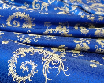 May include: Close-up of a royal blue fabric with a gold floral pattern. The fabric is folded, revealing the intricate design and texture. The pattern includes swirling leaves and flowers.