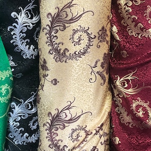 May include: Three rolls of fabric with different patterns. The fabric on the left is black with a silver floral design. The middle fabric is beige with a brown floral design. The fabric on the right is red with a gold floral design.