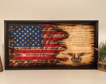 Könnte beinhalten: Eine amerikanische Holzflagge mit einem abgenutzten, verbrannten Look. Die Flagge ist in Schwarz gerahmt und trägt den Text "We The People" aus der US-Verfassung. Die Flagge ist teilweise zerrissen, wobei die Sterne und Streifen sichtbar sind.