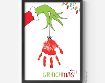 May include: Framed print with a green hand holding a red handprint ornament, with the text "merry GRINCHMAS" in green and red. A green hand in a red sleeve and white cuff is at the top left, with Christmas lights in the upper right.