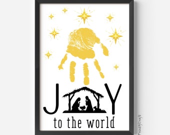 May include: A black framed print featuring a gold handprint and the words "JOY to the world" in black. The handprint is surrounded by gold stars. A black silhouette of the nativity scene is below the handprint.