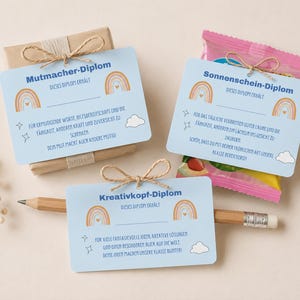 May include: Three light blue rectangular cards with rainbow graphics and German text. One card is tied to a brown paper package, one to a pink snack bag, and one is attached to a wooden pencil with a jute twine bow. Each card features stars and clouds.