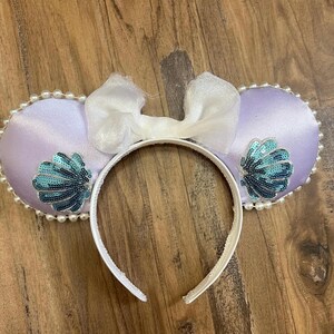 May include: A lavender satin headband with two round ears, each adorned with a seashell design made of teal sequins. The ears are trimmed with faux pearls and topped with a white bow. A fun accessory.