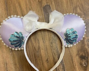 May include: A lavender satin headband with two round ears, each adorned with a seashell design made of teal sequins. The ears are trimmed with faux pearls and topped with a white bow. A fun accessory.