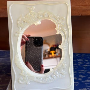 May include: A white plastic vanity mirror with an ornate, raised design around the oval-shaped mirror. The mirror is slightly angled.