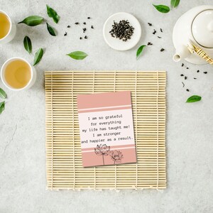 May include: A pink card with a quote about gratitude and happiness. The quote reads: "I am so grateful for everything my life has taught me! I am stronger and happier as a result." The card features a black and white illustration of two lotus flowers.
