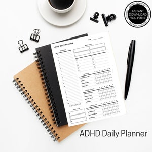 K&ouml;nnte beinhalten: Ein ADHD-Tagesplaner mit einem schwarzen Spiralnotizbuch und einem braunen Spiralnotizbuch. Der wei&szlig;e Planer hat schwarzen Text und enth&auml;lt Abschnitte f&uuml;r die Planung, Tagesziele und Verhaltensverfolgung. Ein schwarzer Stift und eine Kaffeetasse sind ebenfalls sichtbar.