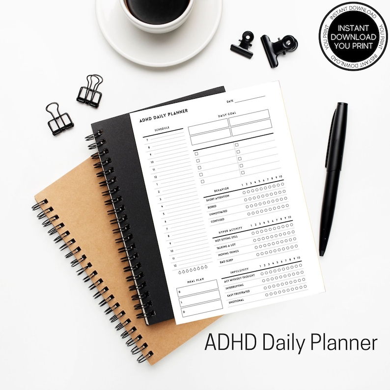 K&ouml;nnte beinhalten: Ein ADHD-Tagesplaner mit einem schwarzen Spiralnotizbuch und einem braunen Spiralnotizbuch. Der wei&szlig;e Planer hat schwarzen Text und enth&auml;lt Abschnitte f&uuml;r die Planung, Tagesziele und Verhaltensverfolgung. Ein schwarzer Stift und eine Kaffeetasse sind ebenfalls sichtbar.