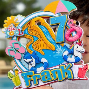 May include: A colorful birthday cake topper with a pool party theme. It features a waterslide, a shark, a flamingo float, and a unicorn float. The number 7 is displayed, along with the name "Frank" and the phrase "at the pool".