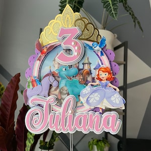May include: A layered cake topper with the name "Juliana" in pink and white script. The topper features a gold crown, the number "3", and various cartoon characters. The background includes a castle scene and a rainbow.