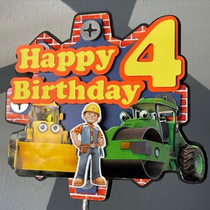 May include: A colorful birthday cake topper featuring the text "Happy Birthday" in yellow and orange, with a large yellow number 4. The design includes cartoon images of construction vehicles and a construction worker.