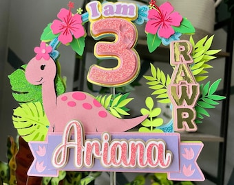 May include: A pink dinosaur-themed cake topper with the text "I am 3" and "Ariana." The topper features pink hibiscus flowers, green leaves, and the word "RAW" in gold. The design is layered and three-dimensional.