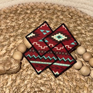 May include: Four square fabric patches with a Southwestern design in shades of red, turquoise, and white. The patches are arranged in a diamond shape, with a black border. The patches are on a woven background with wooden beads.