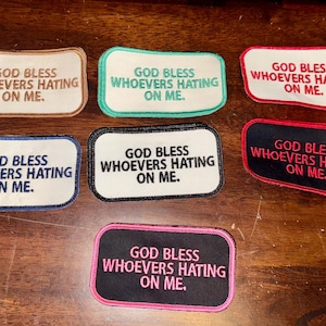 May include: Seven iron-on patches with the text "God Bless Whoever's Hating On Me." The patches are in various colors, including beige, teal, black, white, red, and pink.