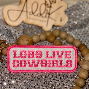 May include: A rectangular patch with the text "LONG LIVE COWGIRLS" in pink embroidery on a white background, framed by a pink border. The patch is set against a sparkly silver background, with wooden beads and a wooden sign in the background.