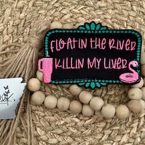 May include: A black and green embroidered patch with the text "Floatin' in the River Killin' My Liver" in pink lettering. A pink flamingo float is in the corner of the patch.