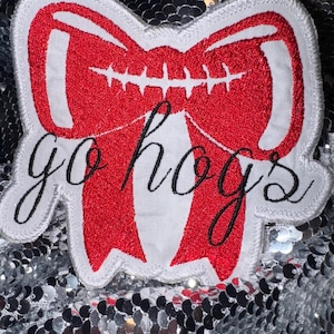 May include: A red and white embroidered patch in the shape of a bow. The bow has a football design in the center and the words "go hogs" in black script. The patch is on a silver sequined background.