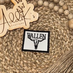 May include: A rectangular patch with the word "WALLEN" above a bull skull graphic. The patch has a black border and is set against a woven background. A wooden sign with the text "Addik's Boutique" and butterfly cutouts is visible.
