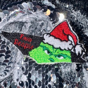 May include: Green Grinch character wearing a red Santa hat with white trim. The Grinch is embroidered on a black and white patch with the text "Grinch People" in red.