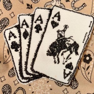 May include: Three white playing cards with black embroidery. The cards are aces and feature a black spade symbol. The centre card shows a black and white embroidered image of a cowboy riding a horse.
