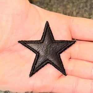 May include: A black star-shaped patch with a textured surface and a stitched border. The patch is designed to be sewn onto clothing or accessories. The star has five points. The patch is made of a dark material.