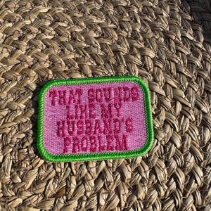 May include: A rectangular patch with the text "THAT SOUNDS LIKE MY HUSBAND'S PROBLEM" in pink lettering. The patch has a pink background with a green border. The patch is set against a woven, light brown background.