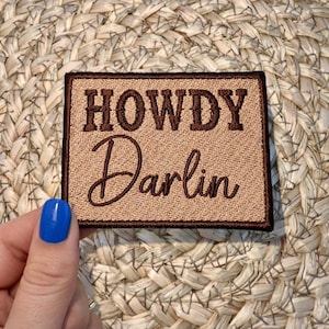 May include: A rectangular patch with the words "HOWDY Darlin" embroidered in brown thread on a tan background. The patch has a brown border and is held by a hand with blue nail polish.