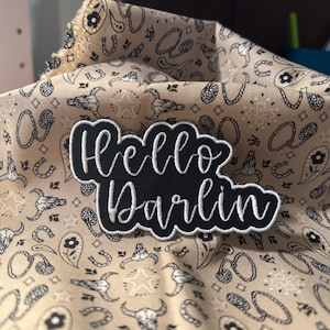 May include: Black and white embroidered patch with the text "Hello Darlin'" on a beige fabric background with a repeating pattern of black and white horseshoes and skulls.