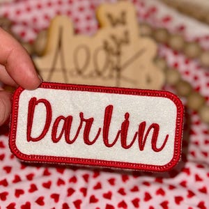 Peut inclure: Un écusson rectangulaire avec le mot "Darlin" brodé en rouge sur un fond blanc. L'écusson a une bordure rouge. Le fond est un tissu avec des cœurs rouges.