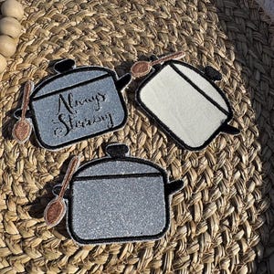 May include: Three embroidered pot-shaped patches with spoons. One patch is gray with the text "Always Stirring". The other two patches are silver and white. All patches have black outlines and small black handles.