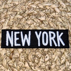 May include: A black rectangular patch with the white embroidered text "NEW YORK" is centered on a woven straw background. The patch is a simple, bold design.