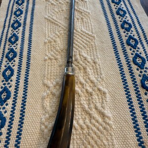 May include: A vintage-style tool with a dark brown handle and a long, slender metal shaft. The handle has a curved shape and a polished finish. The tool rests on a cream-colored woven textile with blue patterned borders.