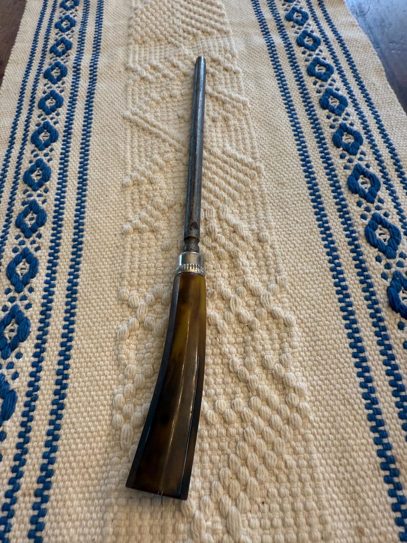 May include: A vintage-style tool with a dark brown handle and a long, slender metal shaft. The handle has a curved shape and a polished finish. The tool rests on a cream-colored woven textile with blue patterned borders.