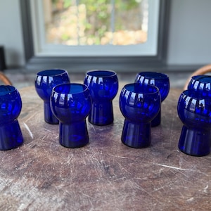 May include: Eight deep blue glass shot glasses with a rounded top and a short, flared base. The glasses are arranged on a wooden surface, reflecting light. These shot glasses are a vibrant addition to any barware collection.