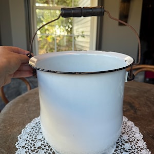 May include: A white enamel bucket with a black rim and a dark wooden handle. The bucket has a wire handle and is sitting on a white lace doily. The bucket is cylindrical and appears to be vintage.