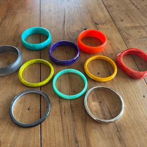 May include: A collection of ten assorted bangle bracelets in various colors including turquoise, orange, purple, yellow, red, lime green, mint green, grey, black, and silver. The bracelets feature different thicknesses and textures.