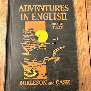 May include: A black book titled "Adventures in English, Grade Three" by Burleson and Cash. The cover features an illustration of a ship sailing on the ocean with a yellow sun in the background.