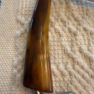 May include: Close-up of a vintage cheese slicer. The handle is a warm, marbled brown and tan color, with a glossy finish. The metal frame is a shiny silver color, with a curved design.