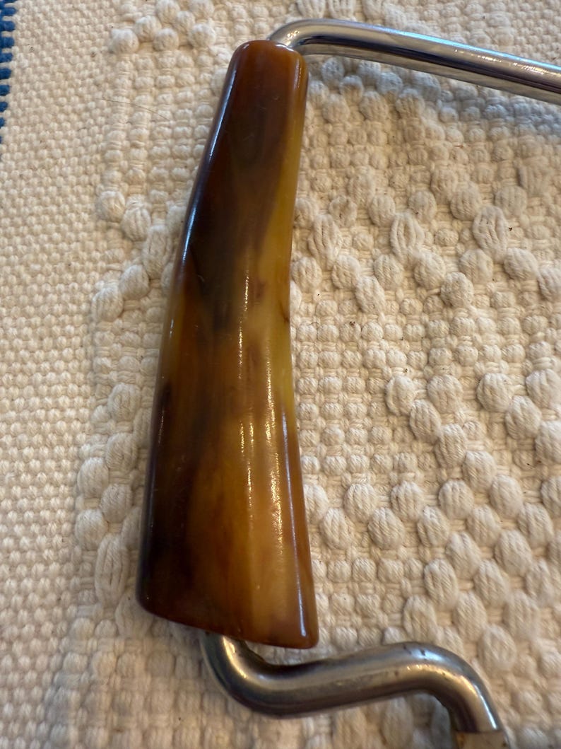 May include: Close-up of a vintage cheese slicer. The handle is a warm, marbled brown and tan color, with a glossy finish. The metal frame is a shiny silver color, with a curved design.
