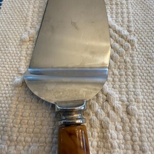 May include: A stainless steel pie server with a brown, marbled handle. The server has a wide, triangular blade and a decorative silver-toned bolster where the handle meets the blade. The handle is a warm, amber color.