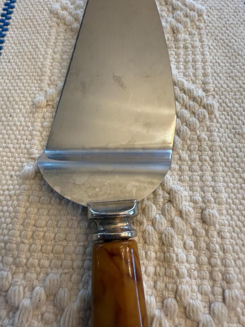 May include: A stainless steel pie server with a brown, marbled handle. The server has a wide, triangular blade and a decorative silver-toned bolster where the handle meets the blade. The handle is a warm, amber color.