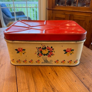 Puede incluir: Panera vintage de metal rojo y blanco con un diseño floral con rosas y una cornucopia. La panera tiene una tapa con bisagras y está en buen estado.