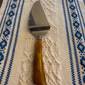 May include: A stainless steel pie server with a brown handle. The server is resting on a cream-colored woven placemat with blue embroidered accents. The server is designed for cutting and serving pies and cakes.