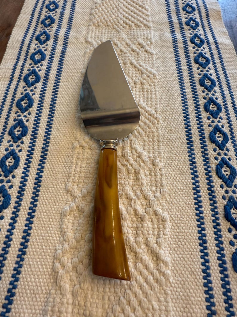 May include: A stainless steel pie server with a brown handle. The server is resting on a cream-colored woven placemat with blue embroidered accents. The server is designed for cutting and serving pies and cakes.