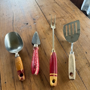Peut inclure: Un ensemble de quatre ustensiles de cuisine comprenant une cuillère à glace en métal avec un manche jaune et rouge usé, un outil pointu en métal avec un manche rouge écaillé, une fourchette à deux dents et une spatule fendue en métal.