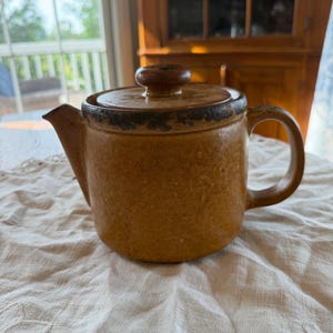 May include: A cylindrical ceramic teapot in a warm, earthy mustard-brown tone with a speckled texture. It features a matching lid with a central knob, a short spout, and a curved handle. The rim of the lid and the upper edge show a darker brown.