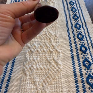 May include: A dark brown bottle stopper with a rounded, oval shape. The stopper is held against a white and blue patterned textile. The textile has a diamond pattern and blue stripes.
