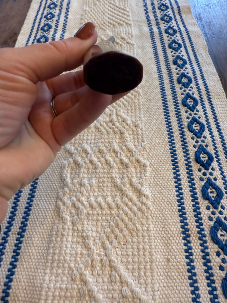 May include: A dark brown bottle stopper with a rounded, oval shape. The stopper is held against a white and blue patterned textile. The textile has a diamond pattern and blue stripes.